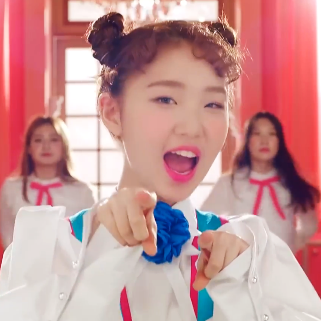 Yeojin from the 'Kiss Later' music video. She has curly brown hair styled into space buns and is wearing a blue dress. She is smiling and pointing at the viewer.